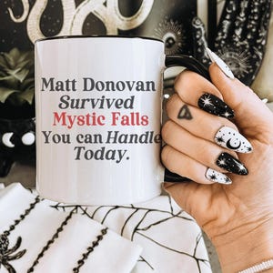 May include: White ceramic coffee mug with a black rim and handle. The mug has black text that reads "Matt Donovan Survived Mystic Falls You can Handle Today."