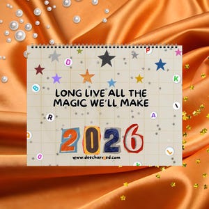 Könnte beinhalten: Ein Spiral-Kalender mit dem Text "LONG LIVE ALL THE MAGIC WE'LL MAKE 2026" und der Website-Adresse www.deecharred.com. Der Kalender ist mit bunten Sternen und Buchstaben auf einem karierten Hintergrund verziert. Der Hintergrund ist ein glänzender, orangefarbener Stoff.