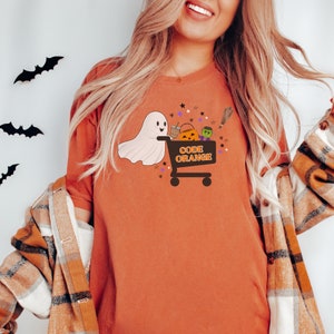 Może przedstawiać: Pomarańczowa koszulka z nadrukiem białego ducha trzymającego wózek zakupowy pełen halloweenowych smakołyków. Na wózku widnieje napis "Kod Pomarańczowy" czarnym kolorem.
