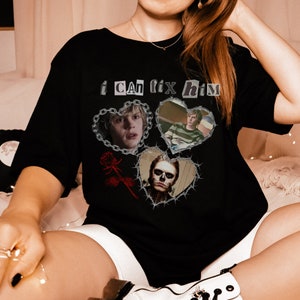Peut inclure: T-shirt noir avec un imprimé graphique de trois cœurs entourés de barbelés. Chaque cœur présente une image différente d'un homme. Le texte "i can fix him" est imprimé au-dessus des cœurs.
