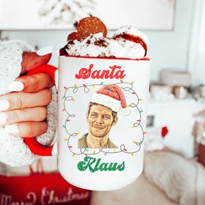 Op de afbeelding: Witte keramische mok met een rode handgreep. De mok heeft een afbeelding van een man met een kerstmanmuts met de tekst "Santa Klaus" in rood en groen.
