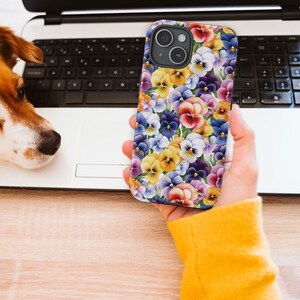 May include: A phone case with a floral pattern featuring purple, yellow, and white pansies. The case is shown on an iPhone 15.
