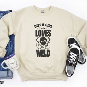 May include: A beige sweatshirt with the text "Just a girl who loves to weld" and a welding mask graphic. The sweatshirt is laid out on a white surface with a pair of black and white sneakers, a pair of blue jeans, and a purple mug with a bee design.