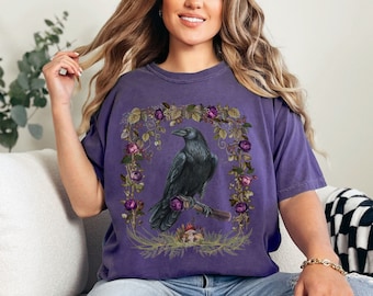 May include: A purple t-shirt with a graphic of a black raven perched on a branch surrounded by purple and green flowers. The raven is in a floral wreath with a skull at the base of the branch.