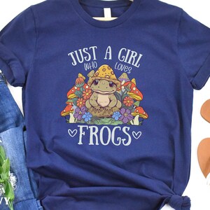 May include: A navy blue t-shirt with the text "Just a girl who loves frogs" in white. A cartoon frog wearing a mushroom hat is surrounded by mushrooms and flowers.