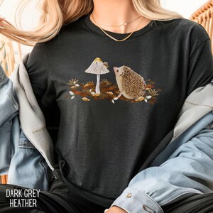 Puede incluir: Una camiseta de color gris oscuro jaspeado con un gr&aacute;fico de un erizo mirando un hongo con un caracol en la parte superior. El hongo es blanco con detalles marrones. El erizo es marr&oacute;n con detalles blancos. El fondo es una escena de bosque marr&oacute;n y amarillo.