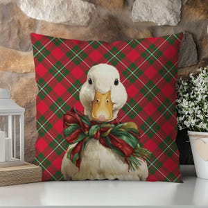 May include: A decorative square pillow featuring a white duck with a yellow beak and a red and green bow. The pillow has a red plaid background with green and white lines. The pillow is a festive home decor item.