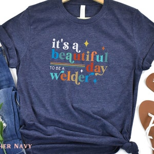 May include: A heather navy blue t-shirt with the text "it's a beautiful day to be a welder" in colorful retro lettering.