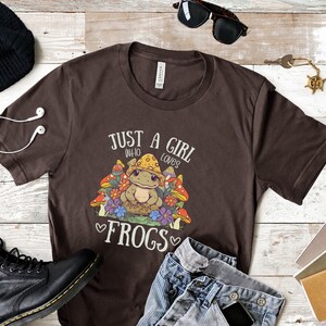 May include: Brown t-shirt with a graphic of a frog sitting in a bed of mushrooms and flowers. The text "Just a girl who loves frogs" is printed on the shirt.