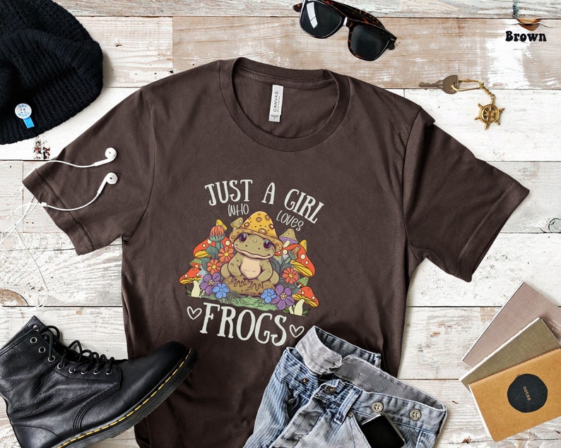 May include: Brown t-shirt with a graphic of a frog sitting in a bed of mushrooms and flowers. The text "Just a girl who loves frogs" is printed on the shirt.