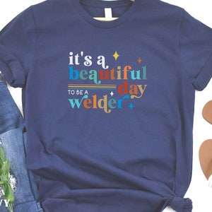 May include: Navy blue t-shirt with the text "it's a beautiful day to be a welder" in colorful letters. The text is decorated with stars and stripes.
