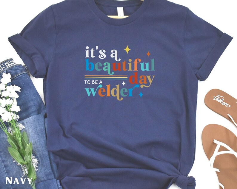 May include: Navy blue t-shirt with the text "it's a beautiful day to be a welder" in colorful letters. The text is decorated with stars and stripes.