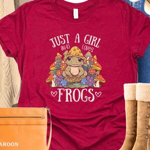 May include: Red t-shirt with a cartoon frog wearing a mushroom hat and surrounded by mushrooms and flowers. The text "Just a girl who loves frogs" is printed on the shirt.