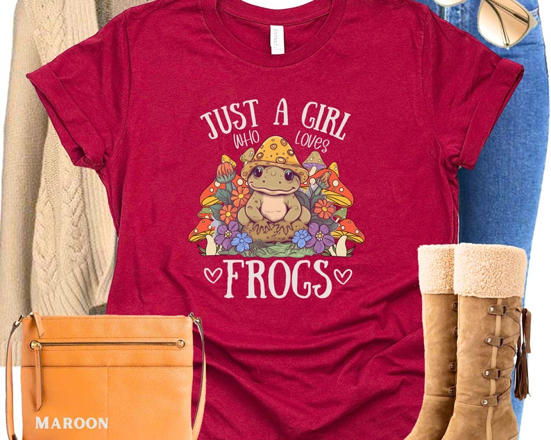May include: Red t-shirt with a cartoon frog wearing a mushroom hat and surrounded by mushrooms and flowers. The text "Just a girl who loves frogs" is printed on the shirt.