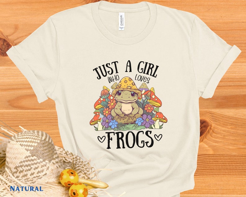 May include: A white t-shirt with the text "Just a girl who loves frogs" in black. The design features a cartoon frog surrounded by mushrooms and flowers.