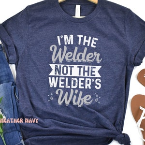 K&ouml;nnte beinhalten: Ein heidefarbenes marineblaues T-Shirt mit dem Aufdruck "I'm the Welder not the Welder's Wife" in Wei&szlig;.