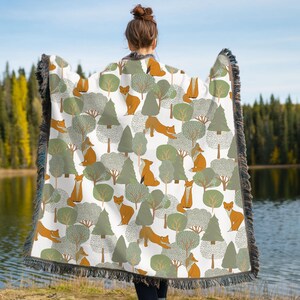 May include: A white woven throw blanket featuring a repeating pattern of orange foxes, green pine trees, and stylized round trees with textured foliage. The blanket has a dark fringed border along all four edges, creating a decorative finish.