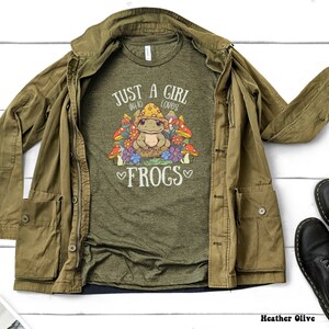 May include: A green t-shirt with the text "Just a girl who loves frogs" and a cartoon frog surrounded by mushrooms. The shirt is laid out on a white surface with a brown jacket and black boots.