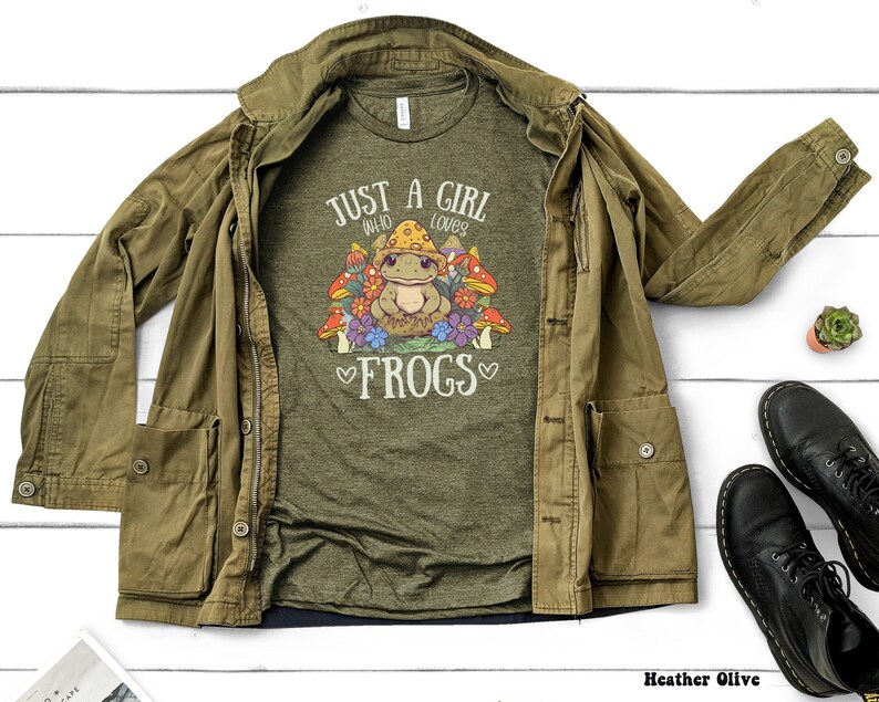 May include: A green t-shirt with the text "Just a girl who loves frogs" and a cartoon frog surrounded by mushrooms. The shirt is laid out on a white surface with a brown jacket and black boots.