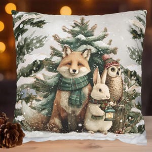 May include: Square decorative pillow featuring a winter scene with a fox, rabbit, and owl. The fox and rabbit wear green scarves, and the owl wears a hat. The background includes snow-covered evergreen trees.