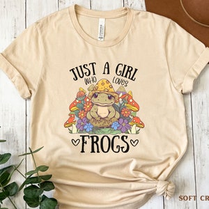 May include: A beige t-shirt with the text "Just a girl who loves frogs" and a cartoon frog wearing a mushroom cap surrounded by mushrooms and flowers.