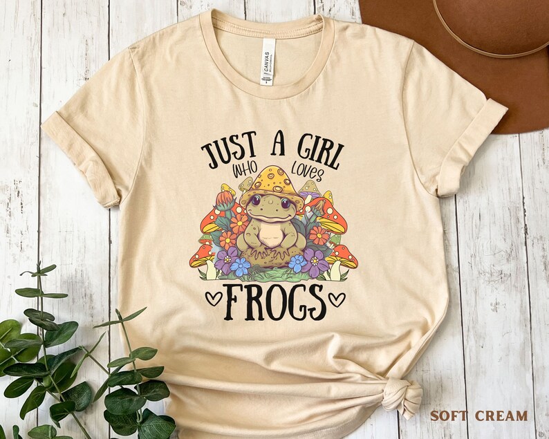 May include: A beige t-shirt with the text "Just a girl who loves frogs" and a cartoon frog wearing a mushroom cap surrounded by mushrooms and flowers.