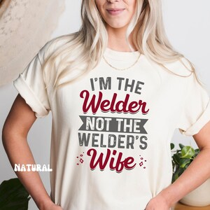 K&ouml;nnte beinhalten: Ein wei&szlig;es T-Shirt mit dem Text "I'm the Welder not the Welder's Wife" in grauer und roter Schrift.
