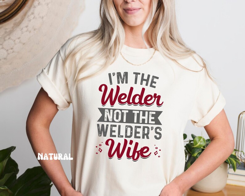 K&ouml;nnte beinhalten: Ein wei&szlig;es T-Shirt mit dem Text "I'm the Welder not the Welder's Wife" in grauer und roter Schrift.