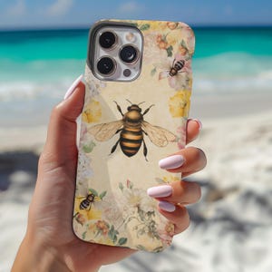 May include: A white iPhone case with a floral design and a bee illustration. The case has a camera lens cutout and a cutout for the phone's buttons.