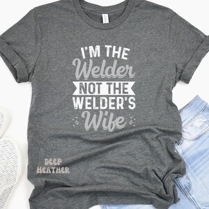 K&ouml;nnte beinhalten: Ein dunkel-heides graues T-Shirt mit dem Aufdruck "I'm the Welder not the Welder's Wife" in wei&szlig;. Das Shirt wird auf einem Model gezeigt, das eine blaue Jeans tr&auml;gt.