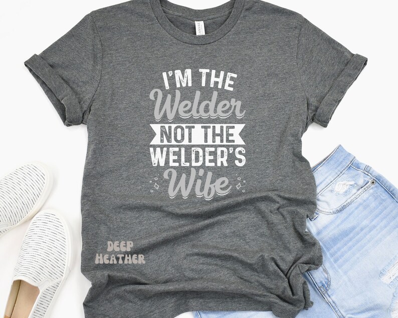 K&ouml;nnte beinhalten: Ein dunkel-heides graues T-Shirt mit dem Aufdruck "I'm the Welder not the Welder's Wife" in wei&szlig;. Das Shirt wird auf einem Model gezeigt, das eine blaue Jeans tr&auml;gt.