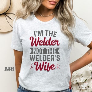 K&ouml;nnte beinhalten: Ein graues T-Shirt mit dem Text "I'm the Welder not the Welder's Wife" in roten Buchstaben.