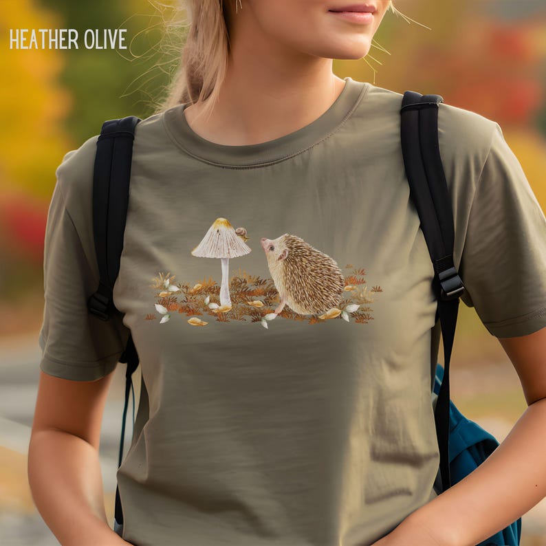 Puede incluir: Una camiseta de color verde oliva jaspeado con un dise&ntilde;o bordado de un erizo mirando a un hongo con un caracol en la parte superior. El dise&ntilde;o est&aacute; en tonos de marr&oacute;n, blanco y verde.