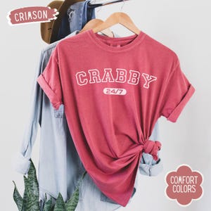 May include: Crimson-colored t-shirt with the word "CRABBY" in white block letters and "24/7" underneath. The shirt is hanging on a wooden hanger with other clothing items in the background. The shirt is knotted at the bottom.