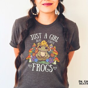 May include: Dark heather grey t-shirt with a cute cartoon frog wearing a mushroom hat and surrounded by mushrooms. The shirt has the text "Just a girl who loves frogs" printed on it.