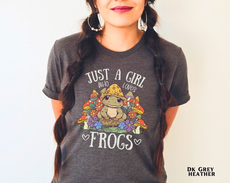 May include: Dark heather grey t-shirt with a cute cartoon frog wearing a mushroom hat and surrounded by mushrooms. The shirt has the text "Just a girl who loves frogs" printed on it.