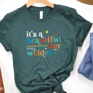 May include: A teal green t-shirt with the text "it's a beautiful day to be a welder" in colorful letters. The text is stylized with a retro font and includes stars.