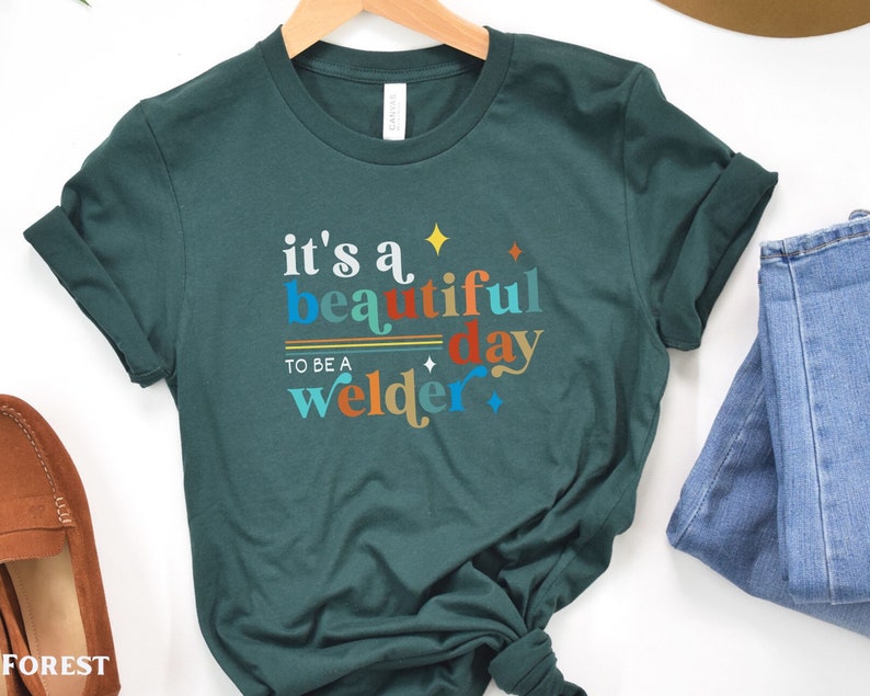 May include: A teal green t-shirt with the text "it's a beautiful day to be a welder" in colorful letters. The text is stylized with a retro font and includes stars.