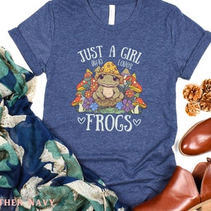 May include: A heather navy blue t-shirt with a graphic of a frog wearing a mushroom hat and surrounded by flowers. The text "Just a girl who loves frogs" is printed on the shirt.