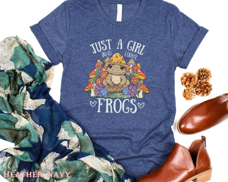 May include: A heather navy blue t-shirt with a graphic of a frog wearing a mushroom hat and surrounded by flowers. The text "Just a girl who loves frogs" is printed on the shirt.
