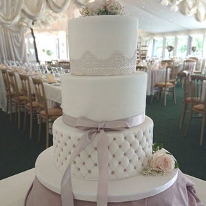 May include: A three-tiered white wedding cake with decorative elements. The top tier features a floral arrangement, the middle tier has a lace design, and the bottom tier has a quilted pattern with pearl accents. A lavender ribbon adorns the cake.