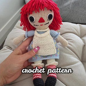 May include: A handmade crochet doll with red yarn hair, black button eyes, and a stitched smile. The doll wears a blue dress, a white apron, and red and white striped socks. The text "crochet pattern" is displayed on the image.