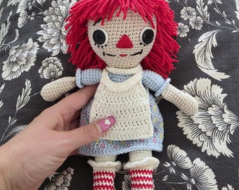May include: A handmade crochet doll with red yarn hair, black button eyes, and a red triangle nose. The doll wears a blue dress, white apron, and red and white striped socks. The doll is a classic toy.