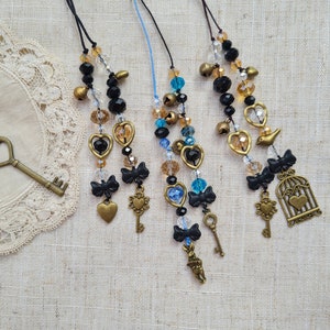 Puede incluir: Cuatro collares de cuentas con dijes, incluyendo llaves, corazones, pájaros y una jaula. Los collares están hechos con cuentas negras, marrones y azules y tienen un estilo rústico y vintage.
