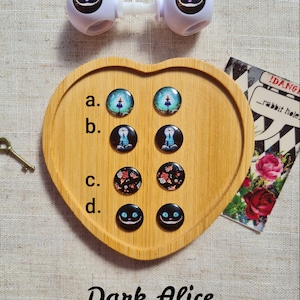 May include: Eight round, colorful, resin buttons with different images from the Alice in Wonderland story. The buttons are arranged in four rows of two on a wooden heart-shaped tray. The tray is on a light brown fabric background. The text "Dark Alice" is written below the tray.