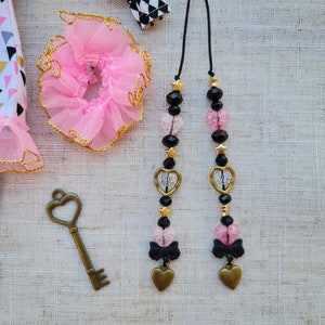 Puede incluir: Dos collares negros y dorados con cuentas de corazón rosa y transparente, estrellas negras y lazos negros. Los collares están sobre un fondo de tela beige.