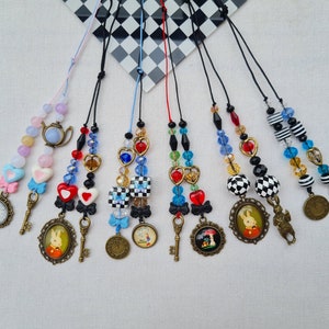 May include: Seven colourful beaded necklaces with charms, including keys, hearts, teapots, and framed images of Alice in Wonderland characters. The necklaces are strung on black cords and feature a variety of beads, including clear, red, blue, and black.