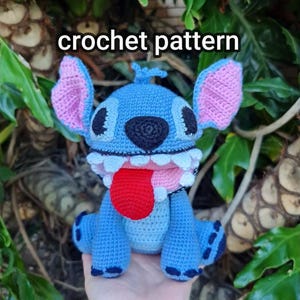 Pode incluir: Um personagem Stitch de crochê azul com orelhas cor-de-rosa, olhos pretos e língua vermelha. O personagem está sentado em uma mão e tem um texto sobreposto "crochet pattern".