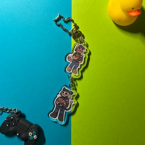 May include: Two clear acrylic keychains with cartoon characters. The keychain on the top features a brown-haired character wearing a green shirt and holding a small white object. The keychain on the bottom features a black-haired character wearing a black shirt and holding a small brown object. Both keychains have silver metal rings.