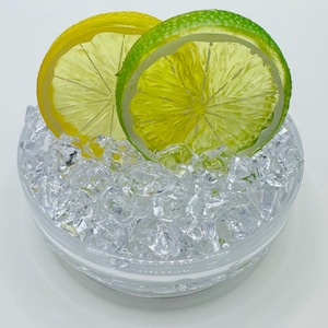 Pu&ograve; includere: Una ciotola di vetro trasparente riempita con piccoli cubetti di ghiaccio in acrilico trasparenti e irregolari. Due fette di frutta artificiale, un limone giallo brillante e un lime verde vivace, sono posizionate verticalmente, parzialmente immerse.